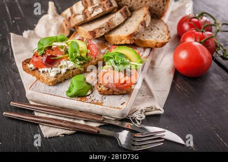 Schließen Sie hausgemachtes Brot mit gerösteten Sandwiches mit Räucherlachs, Avocado, Tomaten, Kräutern und Käse auf einem Holzschneidebrett Stockfoto