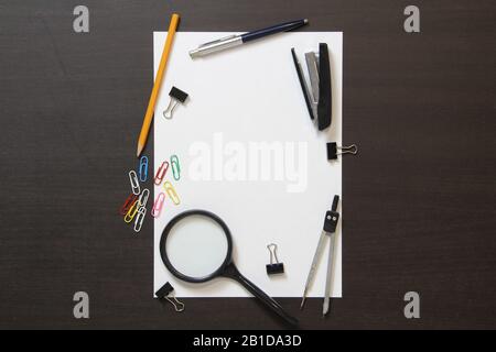 Vorlage für weißes Papier mit Stift, Lupe, Hefter, Büroklammern und anderen Schreibwaren auf dunklem, wengelfarbenem Holzhintergrund. Stockfoto mit leerem Textbereich Stockfoto