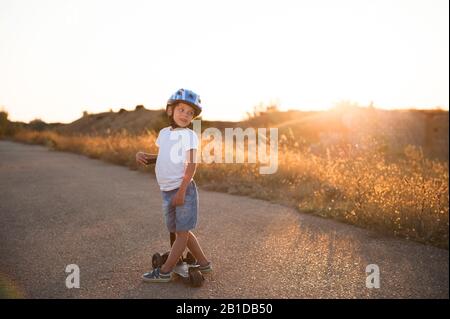 Fröhliches Sport kleines Kind in Helm und blauen Denim-Shorts auf leerem Asphaltweg mit Roller bei sommerlichem warmen Sonnenuntergang Stockfoto