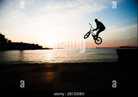 Outdoor-Freizeitsportaktivitäten von jungen Männern, die bei Sonnenuntergang hoch auf dem Extremrad in der Hafenstadt springen Stockfoto