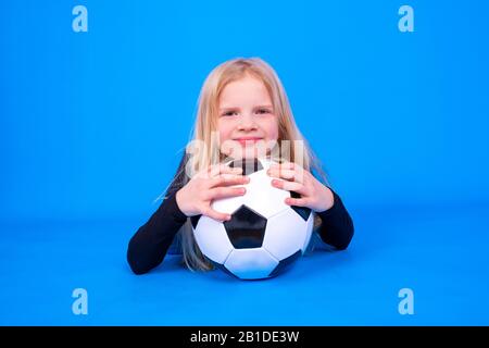 Fußballfans. Süßes blondes Mädchen in schwarzem Hemd, das den Fußballball in Händen über den blauen Studiohintergrund hält. Copyspace. Fußballspiel. Stockfoto