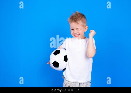 Fußballfans. Fröhlicher kaukasischer Kinderjunge, der den Sieg feiert und den Fußballball in Händen über den blauen Studiohintergrund hält. Copyspace. Fußballspiel. Stockfoto