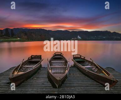 Bled, Slowenien - Traditionelle Boote am Bleder See mit wunderschönem, dramatischem Sonnenuntergang und der Maria-Pilgerkirche und den Bergen am Rücken Stockfoto