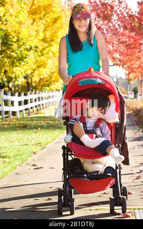 Ältere Teenager-Schwester, die im Herbst behinderten kleinen Jungen in roten Kinderwagen ins Freie drängt Stockfoto