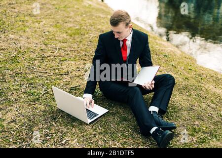 Der junge Student studiert im Park am See die Natur, ein Typ in schwarzem Anzug und roter Krawatte liest einen Laptop und schreibt in einem Notizbuch Stockfoto
