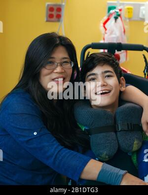 Asiatische Mutter mit Armen um behinderten jungen Teenager-Sohn im Rollstuhl im Krankenhaus, lächelnd zusammen Stockfoto