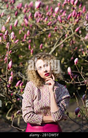 Porträt eines blonden schönen Mädchens mit lockig langen Haaren. Frau spaziert im Garten der blühenden rosafarbenen magnolie Stockfoto