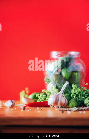 Gurken in Dosen beizen. Zutaten für marinierte Gherkins, Glasbecher, Knoblauch, Dill, Salz, Pfeffer auf rotem Grund. Kopierbereich. Konzept der Gemüsekonservierung Stockfoto