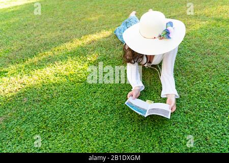 Frau, die sich an einem warmen sonnigen Tag erholt, auf Gras legt und durch den Touristenführer schaut, um das Reiseziel für einen Urlaub zu bestimmen. Reisekonzept. Stockfoto