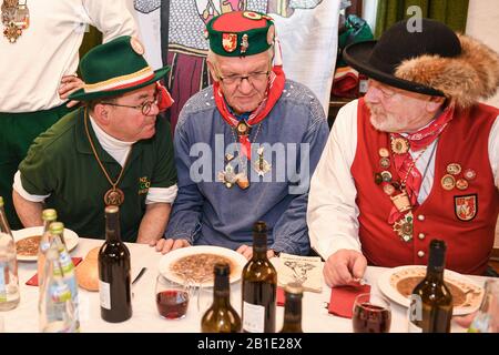 25. Februar 2020, Baden-Württemberg, Riedlingen: Winfried Kretschmann (Bündnis 90/die Grünen, M), Ministerpräsident von Baden-Württemberg, sitzt mit anderen Narren im Rathaus vor seiner Froschtrippe am Schrove-Dienstag. Neben ihnen sitzen Vize-Zunftmeister Lothar Sauter (l) und Ehrenzunftmeister Peter Bucher (r). Mehr als 300 Narren der Narrenzunft Gole 1865 nehmen am Zigarrenrauchen Teil, die 191. Froschtrippe isst und die folgende Rutsche aus dem Rathaus und will später durch die Stadt ziehen. Froschtrippe ist eine Riedlinger Spezialität. Es handelt sich um geschnittene, in Essig eingelegte Rinderstomachen. P Stockfoto