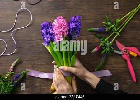Blumenhändler bei der Arbeit: Frau macht Mode modernes Blumenstrauß verschiedener Blumen auf Holzhintergrund. Masterclass. Geschenk für die Hochzeit, Muttertag, Frauentag. Romantische Frühlingsmode. Gefühlvoll violett. Stockfoto