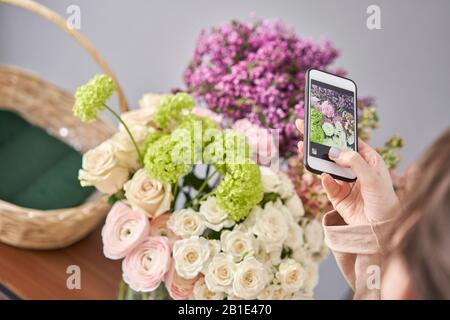 Eine Frau macht ein Foto auf ihrem Telefon mit Blumen. Blumengeschäft Konzept . Blumenfrau schafft Blumenarrangement in einem Korbkorb. . Blumen Lieferung. Stockfoto