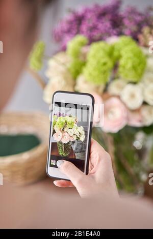 Eine Frau macht ein Foto auf ihrem Telefon mit Blumen. Blumengeschäft Konzept . Blumenfrau schafft Blumenarrangement in einem Korbkorb. . Blumen Lieferung. Stockfoto