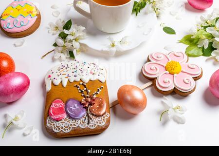 Nahestehende festliche Easter Lebkuchen wie Osterkuchen, Blume und lustige Eier und farbige Eier mit Tasse Tee und weißen Apfelbaumzweigen auf weißem Rücken Stockfoto