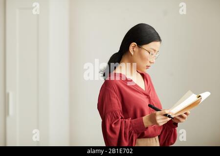 Seitenansicht Porträt erfolgreicher asiatischer Geschäftsfrau mit roter Bluse Lesescheine in der Zwischenablage, während sie im Büro gegen die weiße Wand steht, Kopierraum Stockfoto