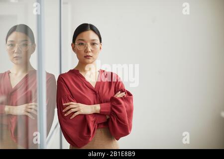 Taillen-Up-Portrait erfolgreicher asiatischer Geschäftsfrau, die selbstbewusst mit den Armen steht, die von der Glaswand im Büro gekreuzt werden, befähigte Frau mit Spiegelung, Kopierraum Stockfoto
