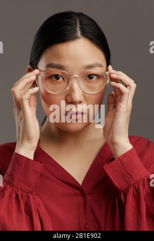 Kopf- und Schulterporträt der eleganten asiatischen Frau, die im Studio vor grauem Hintergrund auf eine Brille steht Stockfoto