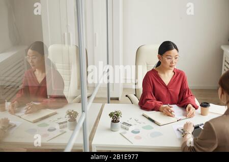 Hochwinkelige Ansicht der modernen asiatischen Geschäftsfrau, die mit dem Kunden spricht, während sie am Schreibtisch in einer weißen Bürozelle, im Kopierbereich, arbeitet Stockfoto