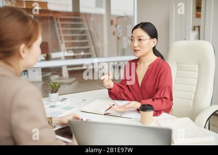 Portrait der lächelnden asiatischen Geschäftsfrau, die mit dem Kunden spricht, während sie am Schreibtisch in der Bürozelle arbeitet, Kopierraum Stockfoto
