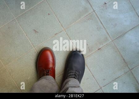 Das Obermaterial aus zusammenpassenden Schuhen, ein Mann, der zwei verschiedene Schuhe und verschiedene Farben auf gefliestem Boden trägt, bricht die Regeln, revolutioniert die Metaphik Stockfoto