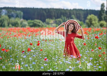 Verträumte Frau im roten Kleid und einem grossen Rot gestreifte Hut in der schönen Kraut blühenden Mohnfeld. Vintage elegant romantisch aussehen. Konzept der schöne Sommer Stockfoto