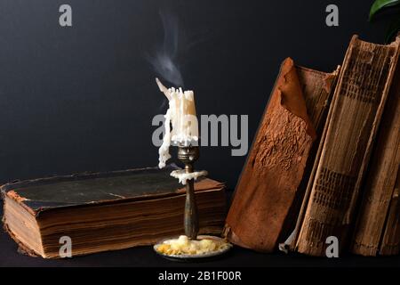 Stapel alter Bücher mit vergilbten Schabbseiten und Kerzenleuchter mit dampfender Kerze Stockfoto