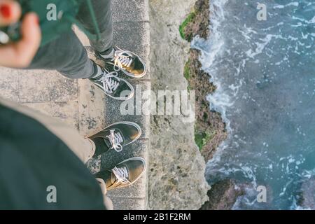 Paar steht neben dem Meer mit passenden Schuhen und abstürzenden Wellen auf Felsen Stockfoto