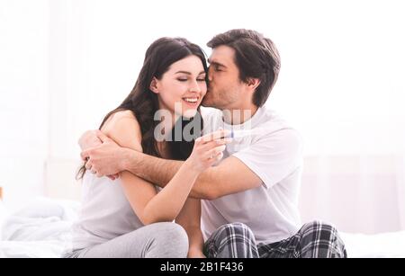 Glücklicher Ehemann, der seine Frau mit positivem Schwangerschaftstest küsst Stockfoto