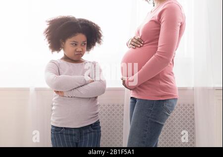 Beleidigte kleine schwarze Tochter, die mit Eifersucht auf den Bauch der schwangeren Mutter blickt Stockfoto