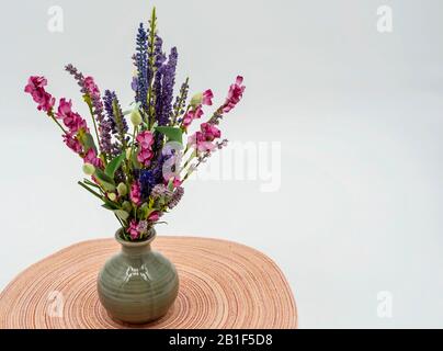 Pink und Purple künstliche Wildblumen in einer grauen Keramikvase, die auf einem runden rosafarbenen und weißen Schild mit weißem Hintergrund sitzt. Stockfoto