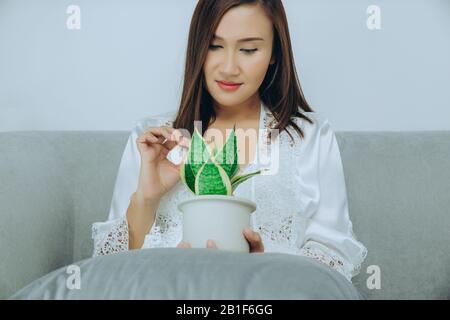 Asiatische Frauen tragen weiße Nachtgown- und langärmlige Satin-Robe mit Blumenspitze halten Luftreinigungsanlagen, die auf dem grauen Sofa im Schlafzimmer bei nig sitzen Stockfoto