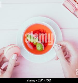 Weibliche Hände, die eine weiße Tasse Himbeertee und einen pinkfarbenen Makronen auf einem blassrosa Hintergrund halten. Draufsicht Stockfoto