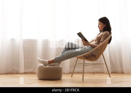 Entspanntes Mädchen liest Buch im Stuhl am Fenster
