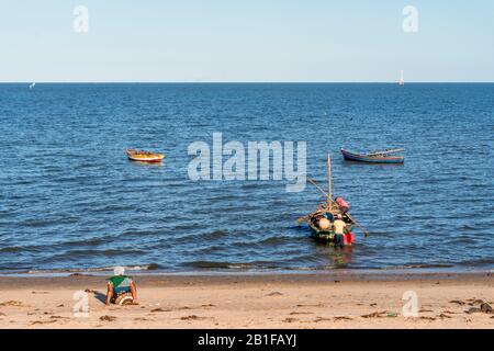 Ein Fischer, der an seinem Boot arbeitet, während eine Frau am Strand sitzt, Costa do Sol, Maputo, Mosambik Stockfoto