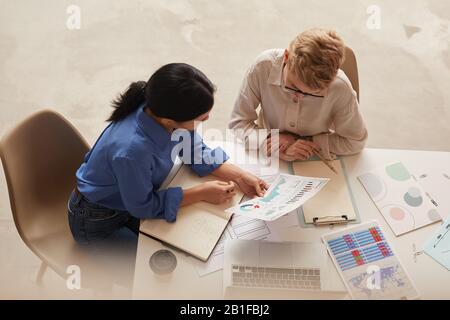 Hochwinkelansicht bei zwei modernen Geschäftsfrauen, die Datendiagramme diskutieren, während sie am Tisch sitzen und Platz für Kopien schaffen Stockfoto