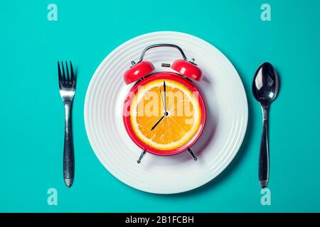 Roter Wecker mit orangefarbenem Obst statt Uhrenrad auf dem weißen Teller mit Löffel und Gabel auf dem Farbhintergrund Stockfoto