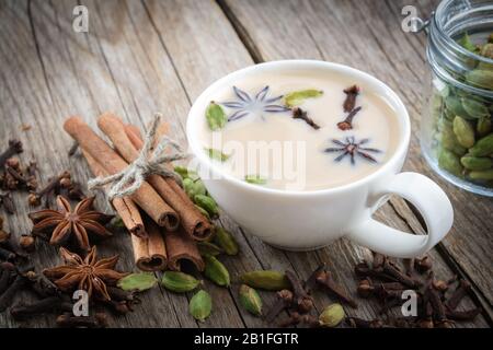 Tasse gesunden ayurvedischen Masala-Tee oder Kaffee mit Milch und aromatischen Gewürzen. Zimtstangen, Kardamom-Schoten, Nelken und Anissterne auf Holztisch. Stockfoto