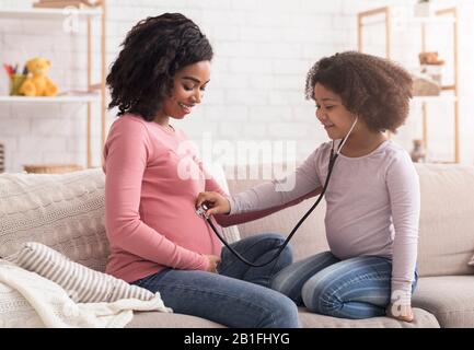 Kleines schwarzes Mädchen, das schwangeren Bauch der Mutter mit Stethoskop hört, spielender Arzt Stockfoto