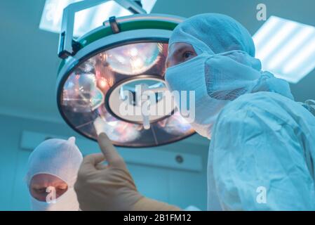 Nahaufnahme von Chirurgen in einer sterilen Maske, vor dem Hintergrund einer Operationslampe, mit erhobenen Händen, der Assistent des Chirurgen sieht den Indikator an Stockfoto