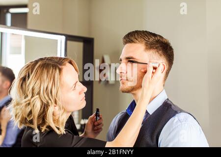 Vorbereitung des professionellen Schauspielers vor dem Dreh. Gutaussehender junger Mann, der sich von einem professionellen visagisten bewirbt. Stockfoto