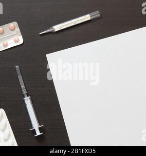 Vorlage für weißes Papier mit Pillen in Blister, Thermometer und Spritze zur Injektion auf dunklem, wengem Holzhintergrund. Die Bereitstellung medizinischer und pharmakologischer Dienstleistungen. Stockfoto