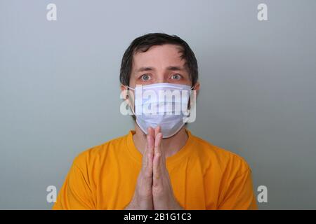 Erwachsene Männer mittleren Alters in einem gelben T-Shirt auf grauem Hintergrund in einer medizinischen Maske halten seine im Gebet gefalteten Handflächen fest. Krankheitsschutz Coronavirus epidemisches Problem aus China. Stockfoto