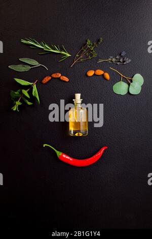 Lächelndes Gesicht aus Kräutern und Flasche ätherisches Öl auf schwarzem Hintergrund, Draufsicht, flacher Lay Stockfoto