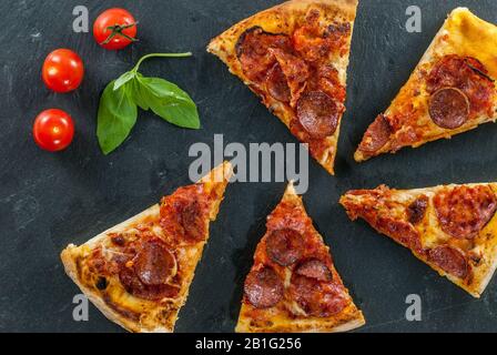 Köstliche frisch gekochte Scheiben von Pfefferoni Pizza auf Schiefer mit Kirschtomaten, rotem Pfeffer und Basilikumblättern Stockfoto