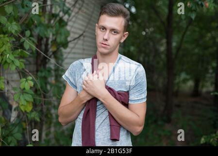 Porträt eines jungen Kerls, der mit gekreuzten Armen steht. In der Nähe des alten Gebäudes steht ein Mann mit rotem Pullover, an der Wand ein Wildivy. Stockfoto