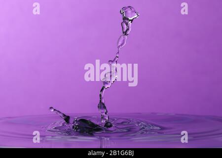 Abstraktes Foto einer Wasserfallenkollision, die mit zwei Wassertropfen erzeugt wurde, die isoliert vor einem violetten Hintergrund zusammenspritzen. Stockfoto