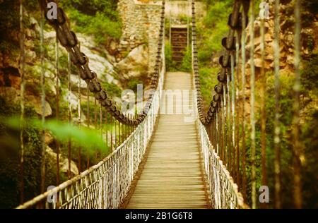 Gefederte Fußgängerbrücke über den Fluss, selektiver Fokus auf den Vordergrund. Felsen von Pflanzen im Hintergrund. Stockfoto