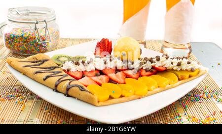 Mit Erdbeer- und Pfirsichscheiben garniert mit Schokolade, Sahne und Frucht-Eis auf einem weißen Teller mit zwei Glaskalen mit Saft auf weißer BA Stockfoto