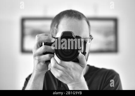 Selbstporträt durch einen Spiegel eines nicht erkennbaren kaukasischen Mannfotografen, der eine Kamera hält (Fokus auf Kameralinse). Stockfoto