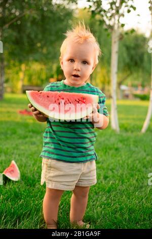 Süßer kleiner Junge, der große Slice-Wassermelone im Garten isst. Blondes Kind, das bei einem Picknick im Sommer bei Sonnenuntergang Wassermelonen im Freien isst. Gesunde Früchte Stockfoto